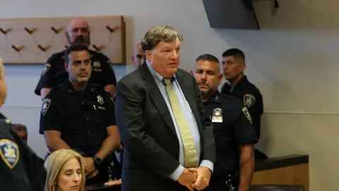 Gilgo Serial Killer Rex Heuermann stands in a dark jack, white shirt and yellow tie. He is speaking in a courtroom, and there are several police in uniform around him. A woman is sitting at a table next to him.